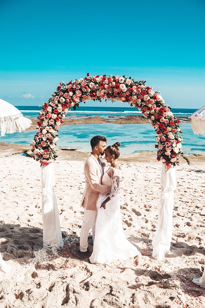 Hochzeitspaar steht am Strand unter einem Traubogen, während der Bräutigam die Braut auf die Stirn küsst